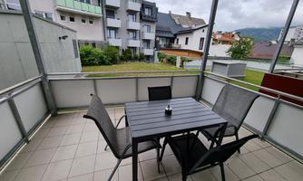 A cozy balcony with a black table and four chairs. In the background, buildings and a green garden are visible.