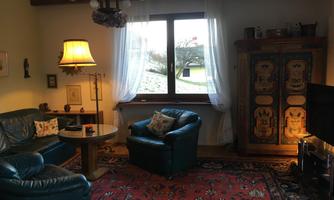 A cozy living room with blue leather furniture and a round table. White curtains hang by a window, and in the background, there is a colorful cupboard.