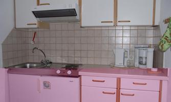 A small kitchen with a pink countertop and white cabinets. On the counter, there is a kettle and a coffee maker.