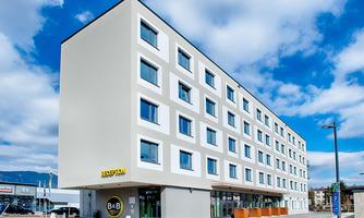A modern hotel building with five floors and large windows. The facade is bright and simple, surrounded by a clear sky.