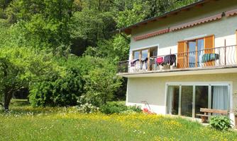 A house with a balcony on which laundry is hanging, surrounded by a green garden. In the background, trees and a natural landscape are visible.