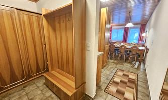 A cozy entrance area with wood paneling and a coat rack. In the background, a dining area with a large table and chairs is visible.