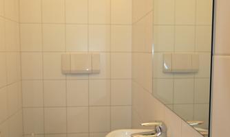 A simple bathroom with white tiles, a toilet, and a small sink. The space is bright and modernly designed.