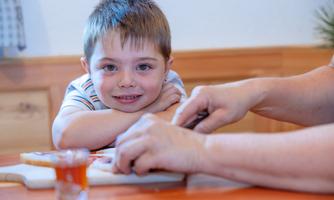 A smiling boy is watching attentively as a hand prepares a sandwich for him. On the table, there is a small glass with a drink.