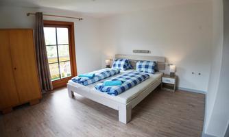 A bright bedroom with a double bed and striped bed covers. There is a large window with a view outside and a wooden wardrobe.