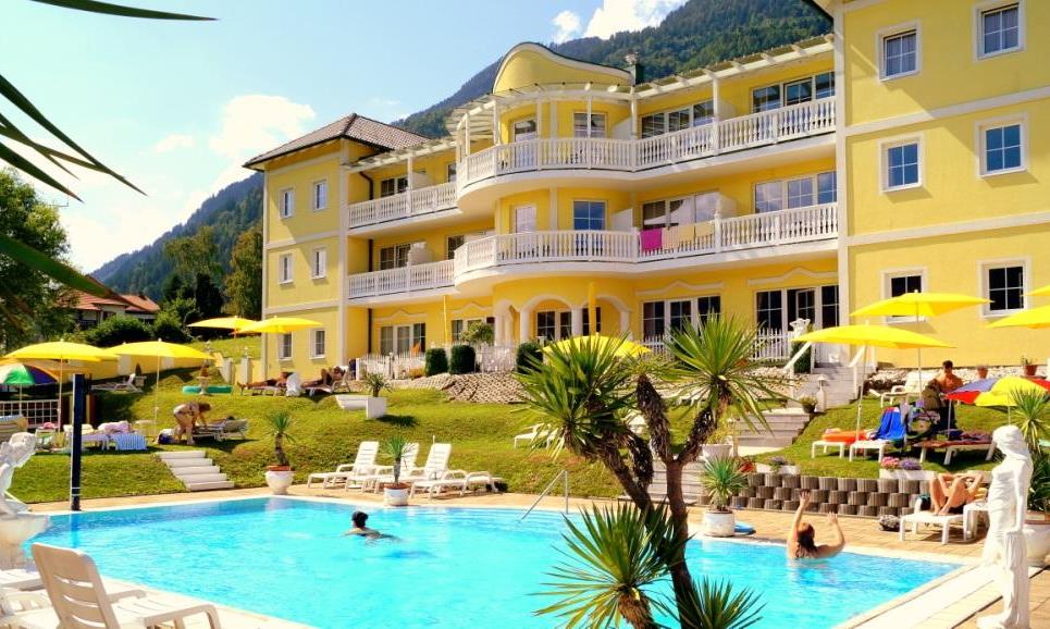 An elegant hotel with a yellow facade, surrounded by green mountains. The outdoor pool invites relaxation, while lounge chairs and umbrellas are available.