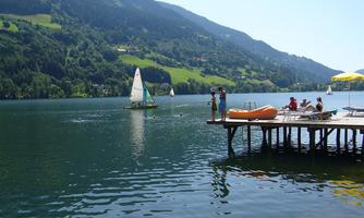 A beautiful lake with a sailing boat and green mountains in the background. People are relaxing on a pier and enjoying the sunny weather.