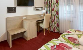 A cozy hotel room with a double bed and colorful floral patterns. There is a desk, a television, and large windows with curtains.