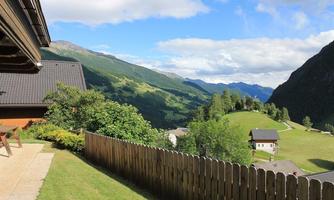 A picturesque landscape with mountains and a green meadow. In the foreground, a wooden fence and a cozy house can be seen.