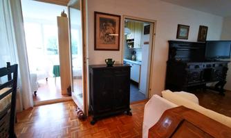 A bright room with wooden flooring and an open connection to the kitchen. The ceiling and walls are simple, and there are some pieces of furniture, including a black wardrobe.