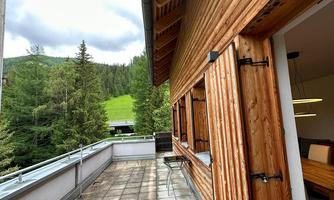 A terrace with tiles and wood paneling, surrounded by trees and green meadows. The sky is partly cloudy, creating a calm atmosphere.