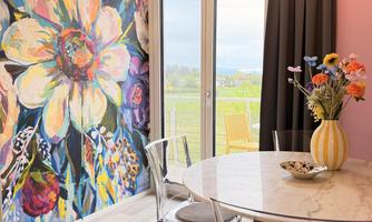 A brightly decorated room with a colorful flower mural and a round table. Through the large windows, one looks out onto a green landscape.