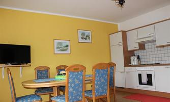 A cozy dining area with a round table and blue chairs. The yellow wall and the kitchen unit give the room a friendly atmosphere.