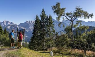 Two hikers are standing at a signpost in the mountains. The view extends over a wooded landscape with high mountains in the background.