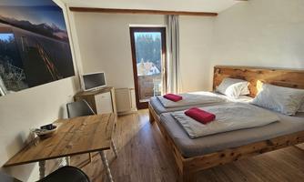 A cozy room with two beds and a wooden desk. Large windows let in a lot of daylight and offer a view of the surroundings.