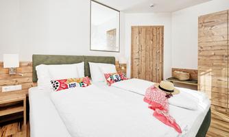 A modern bedroom with a large double bed and light wood decor. On the bed are a hat and a colorful scarf.