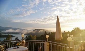 A terrace with a table and chairs overlooks a picturesque lake. The morning sun shines through the clouds and illuminates the landscape.