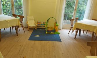 A bright room with wooden floors and several tables. In the center, there is a play mat with toys for toddlers.