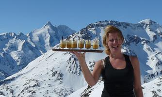 A woman is holding a tray with drinks in the mountains. In the background, snow-capped peaks and a blue sky can be seen.