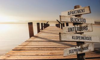 A wooden walkway leads over the water with several signposts. The signs indicate directions to various places such as "Apartment" and "Klopeinersee".