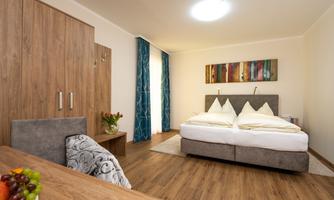 A modern bedroom with a large double bed and wooden floor. Colorful pictures hang on the wall, and there is a small table with fruit.