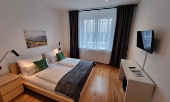 A modern bedroom with a double bed and wooden floor. There is a television and large windows with curtains.
