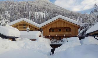 Two log cabins in a snowy landscape with high mountains in the background. A dog is running through the snow between the houses.