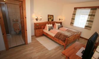 A cozy bedroom with a double bed and wooden furniture. In the background, there is a bathroom with a shower.