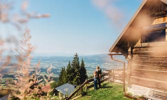 A beautiful wooden house in nature with a view of the valley. Two people are standing in the meadow, surrounded by trees and grass.