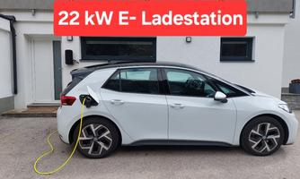 A white electric car is charging at a 22 kW E-charging station. In the background, a modern building can be seen.