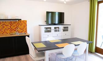 A modern kitchen with a dining area and a large television. The walls are light, and the space has colorful accents.