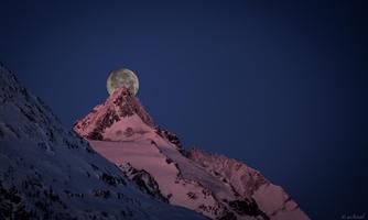 A majestic mountain peak covered in snow, with the full moon sitting on it. The landscape is bathed in soft evening light.