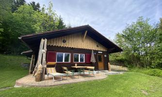 A cozy wooden house amidst green grass and trees. Outside, garden furniture is placed on a wooden terrace.