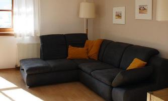 A cozy living room with a large, dark sofa and orange cushions. There is a table and pictures on the wall, as well as plenty of natural light.