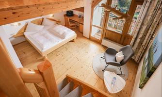 A cozy bedroom with wooden decor and large windows. There is a double bed and a modern armchair on a round rug.