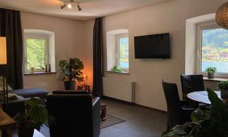 A cozy living space with a television and a dining table. Large windows offer views of the water and the surroundings.