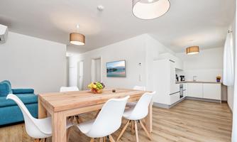 A modern living room with a large wooden table and white chairs. The kitchen is bright and well-equipped, with ample space for cooking and eating.