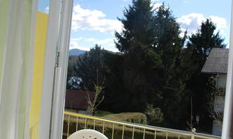 A small balcony with two white chairs and a round table. In the background, green trees and a blue sky with some clouds can be seen.