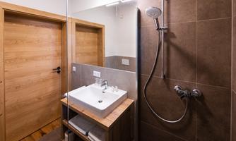 A modern bathroom with a shower and a sink on a wooden frame. The walls are in neutral brown tones and a wooden door is visible.