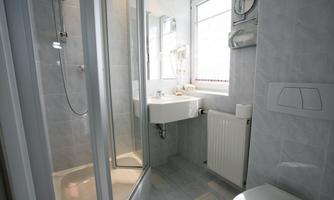 A modern bathroom with a shower, a sink, and a toilet. The walls are light tiled and the window provides daylight.