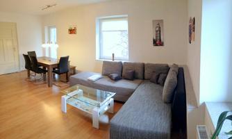 A modern living room with a gray sofa and a glass table. There is a dining table with chairs and large windows that let in plenty of light.