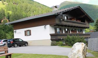 A modern house in the mountains with a parking space and a beautiful green meadow. In the background, gentle hills and trees can be seen.