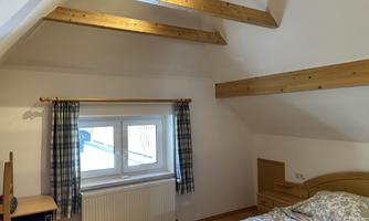 A bright bedroom with wooden ceiling beams and a large window. The furnishings consist of a bed and a small desk.