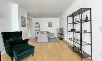 A modern living room with a green armchair and a sofa. An elegant shelf houses decorative items and the floor is made of wood.