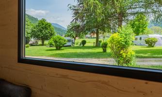 A window view of a green landscape with trees and hills. In the foreground, you can see a cozy living space.