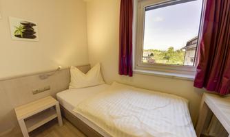 A simple room with a single bed and a window that overlooks a green landscape. The room is bright and modernly furnished.