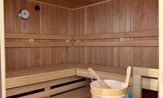 A relaxing sauna area made of wood with benches and a bucket. A clock hangs on the wall, and there is a bottle of liquid on a shelf.