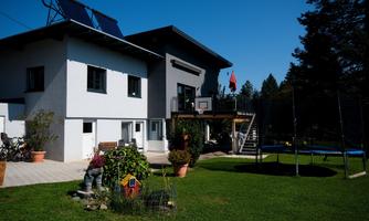 A modern house with solar panels and a balcony. In the garden, there is a playground with a trampoline and beautiful plants.