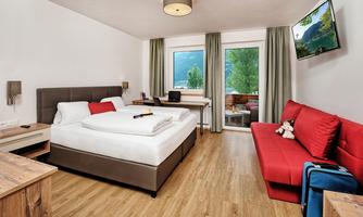 A modern room with a double bed and a red sofa. Large windows offer a view of greenery.