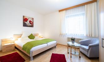 A bright bedroom with a double bed and a cozy armchair. The walls are in neutral colors, and a picture with flowers hangs above.